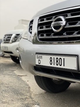 A close-up view of several parked Nissan cars, focusing on the front grille and license plate of the nearest vehicle, which has a Dubai registration number.