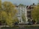 A building under renovation is partially covered with white tarp. The structure features classic European architecture with brick walls and arched windows. In the foreground, trees with yellow-green leaves are present, and various vehicles, including bicycles and vans, are parked along the street.