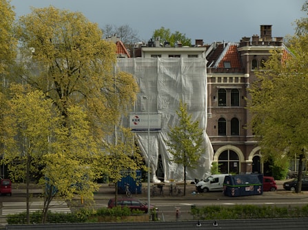 A building under renovation is partially covered with white tarp. The structure features classic European architecture with brick walls and arched windows. In the foreground, trees with yellow-green leaves are present, and various vehicles, including bicycles and vans, are parked along the street.