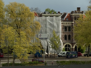 A building under renovation is partially covered with white tarp. The structure features classic European architecture with brick walls and arched windows. In the foreground, trees with yellow-green leaves are present, and various vehicles, including bicycles and vans, are parked along the street.