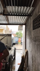 A white vehicle is parked inside a narrow garage with a corrugated metal roof. The side of the vehicle is partially open, and a person is standing next to it on the left side of the image. The garage opens up to an outdoor area where orange traffic cones and additional vehicles are visible. The walls of the garage are plain and the floor appears wet, suggesting it may be raining.