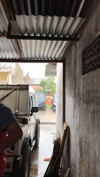 A white vehicle is parked inside a narrow garage with a corrugated metal roof. The side of the vehicle is partially open, and a person is standing next to it on the left side of the image. The garage opens up to an outdoor area where orange traffic cones and additional vehicles are visible. The walls of the garage are plain and the floor appears wet, suggesting it may be raining.