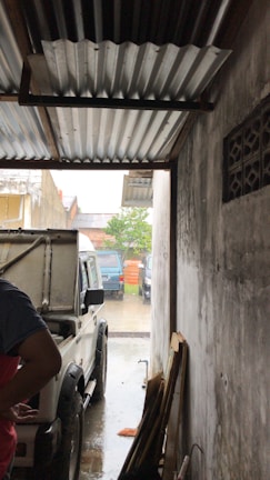A white vehicle is parked inside a narrow garage with a corrugated metal roof. The side of the vehicle is partially open, and a person is standing next to it on the left side of the image. The garage opens up to an outdoor area where orange traffic cones and additional vehicles are visible. The walls of the garage are plain and the floor appears wet, suggesting it may be raining.