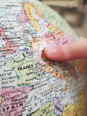 A close-up view of a globe showing the map of Europe with a finger pointing at the area around Switzerland, Italy, and France. The globe displays various country names, cities, and geographical features in colored sections.