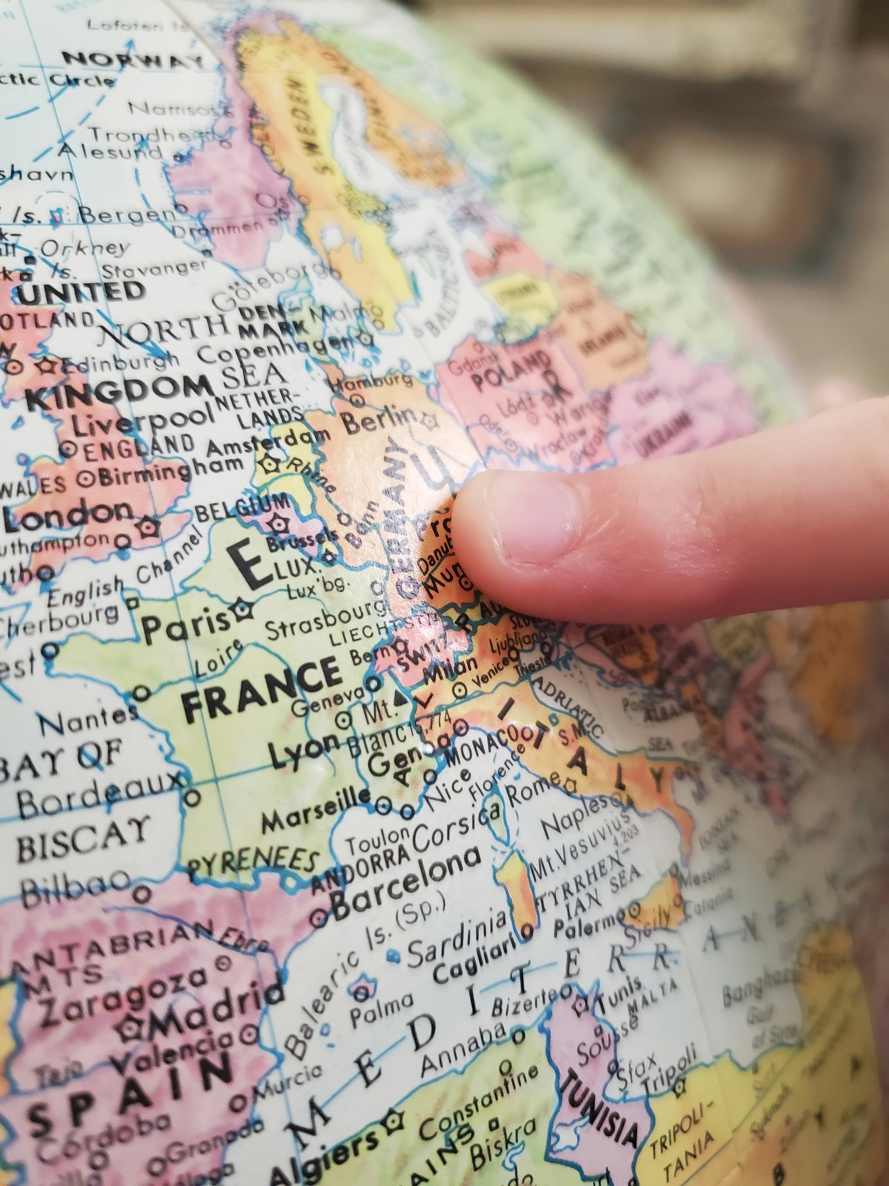 A child's finger points to Germany on a globe. The bright colors of the map has bold black letters indicating places in the world.