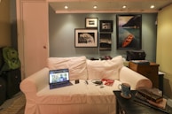 A cozy living room scene with a white sofa placed against a wall decorated with framed photographs and art. On the sofa, there is an open laptop, a smartphone, and earphones. On the right side, a coffee table holds a blue mug, notebooks, and a few small items. To the left, backpacks are stacked next to the sofa, and a small wooden cabinet is visible on the right.
