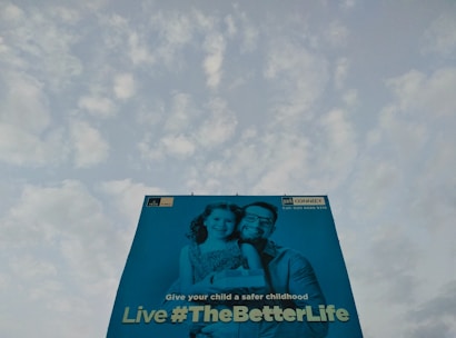 A large billboard features an image of a man and a child smiling, with a message promoting a safe childhood. The background is a clear sky with scattered clouds.