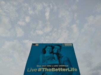 A large billboard features an image of a man and a child smiling, with a message promoting a safe childhood. The background is a clear sky with scattered clouds.