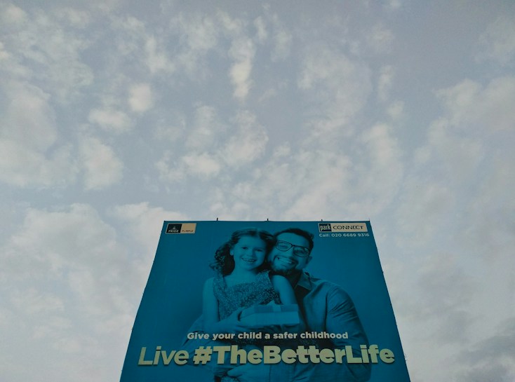 A large billboard features an image of a man and a child smiling, with a message promoting a safe childhood. The background is a clear sky with scattered clouds.