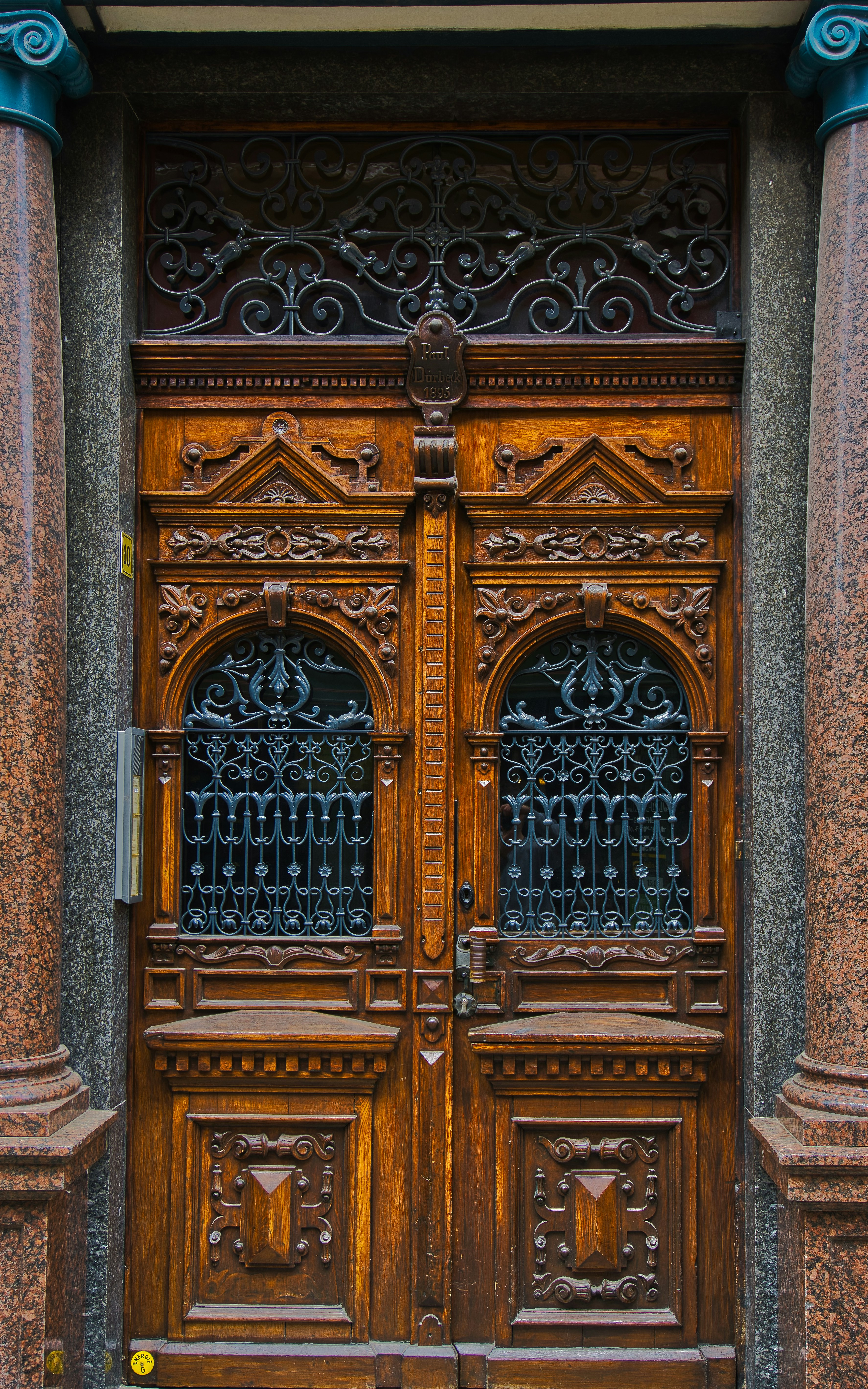 A detailed and ornately carved wooden double door with intricate wrought iron grills in the top half. The wood is rich and dark, flanked by polished stone columns. The doors have a symmetrical design with geometric patterns and ornamental details, conveying a classic and elegant aesthetic.