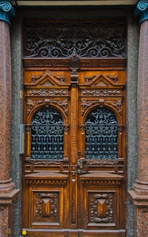 A detailed and ornately carved wooden double door with intricate wrought iron grills in the top half. The wood is rich and dark, flanked by polished stone columns. The doors have a symmetrical design with geometric patterns and ornamental details, conveying a classic and elegant aesthetic.