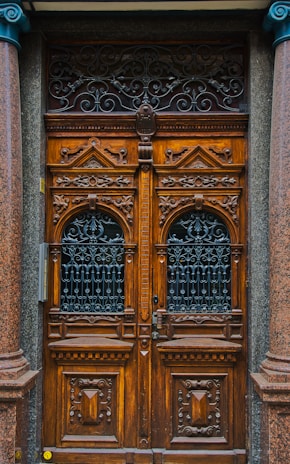 A detailed and ornately carved wooden double door with intricate wrought iron grills in the top half. The wood is rich and dark, flanked by polished stone columns. The doors have a symmetrical design with geometric patterns and ornamental details, conveying a classic and elegant aesthetic.