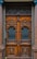 A detailed and ornately carved wooden double door with intricate wrought iron grills in the top half. The wood is rich and dark, flanked by polished stone columns. The doors have a symmetrical design with geometric patterns and ornamental details, conveying a classic and elegant aesthetic.