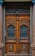 A detailed and ornately carved wooden double door with intricate wrought iron grills in the top half. The wood is rich and dark, flanked by polished stone columns. The doors have a symmetrical design with geometric patterns and ornamental details, conveying a classic and elegant aesthetic.