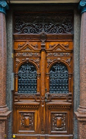 A detailed and ornately carved wooden double door with intricate wrought iron grills in the top half. The wood is rich and dark, flanked by polished stone columns. The doors have a symmetrical design with geometric patterns and ornamental details, conveying a classic and elegant aesthetic.