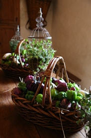 A rustic setting with wicker baskets filled with fresh vegetables like green and purple bell peppers, placed on a wooden table. Decorative wire cages with green plants are positioned behind the baskets, enhancing the natural and organic feel.
