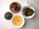 A neatly arranged assortment of items on a white surface includes a bowl of steaming golden tea, a small container with dried tea leaves, a plate with dark chocolate cookies featuring white nuts, and a dish of wet, leafy green tea leaves. The white ceramic teaware adds a refined touch.