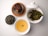 A neatly arranged assortment of items on a white surface includes a bowl of steaming golden tea, a small container with dried tea leaves, a plate with dark chocolate cookies featuring white nuts, and a dish of wet, leafy green tea leaves. The white ceramic teaware adds a refined touch.