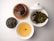 A neatly arranged assortment of items on a white surface includes a bowl of steaming golden tea, a small container with dried tea leaves, a plate with dark chocolate cookies featuring white nuts, and a dish of wet, leafy green tea leaves. The white ceramic teaware adds a refined touch.