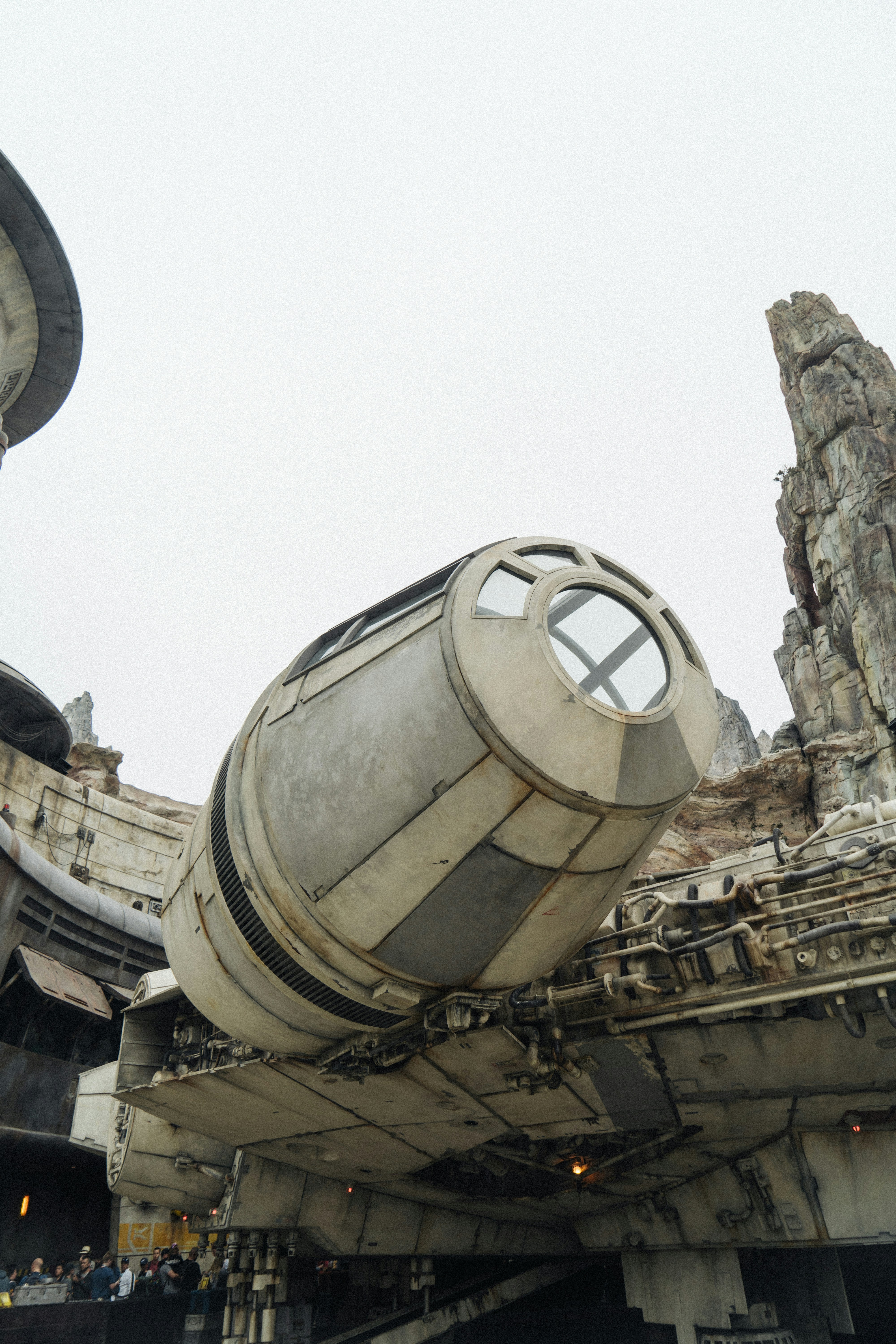 A close-up view of a large, detailed spacecraft model with distinctive round and angular features, resembling a well-known fictional spaceship. The scene is set outdoors, with rocky formations and futuristic architecture in the background. A group of people is visible at the bottom left, adding perspective to the scale of the spacecraft.