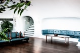 A minimalist interior with white walls and wooden flooring. The room features a blue cushioned seating area arranged along the curved wall, accompanied by a wooden table and a planter with lush green leaves. There are stairs leading to an upper level through an arched opening. Overturned chairs are placed on one of the seats.