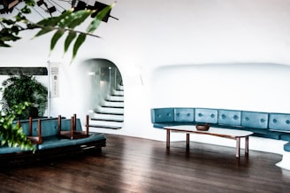 A minimalist interior with white walls and wooden flooring. The room features a blue cushioned seating area arranged along the curved wall, accompanied by a wooden table and a planter with lush green leaves. There are stairs leading to an upper level through an arched opening. Overturned chairs are placed on one of the seats.