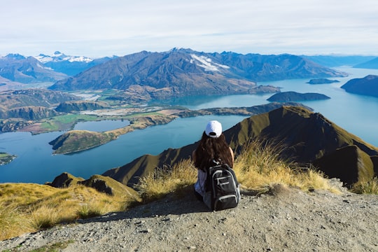 person in black jacket and white helmet sitting on rock looking at mountains during daytime in Roys Peak New Zealand