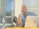 A smiling man with short hair, sitting at a desk with a laptop and a notepad.