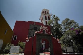A vibrant red and white colonial-style church with ornate architectural details stands prominently against a clear blue sky. The structure features a tall tower with multiple windows and decorative elements. Green trees with dense foliage surround the building, adding a natural backdrop to the scene. Signs with text are posted on the perimeter fence.