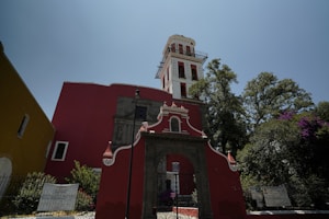 A vibrant red and white colonial-style church with ornate architectural details stands prominently against a clear blue sky. The structure features a tall tower with multiple windows and decorative elements. Green trees with dense foliage surround the building, adding a natural backdrop to the scene. Signs with text are posted on the perimeter fence.