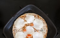 A pizza with toppings of cheese and tomato slices is placed in a black, square pan. The cheese is partially melted, and the pizza seems to be cooking. The surface beneath the pan is dark, possibly a stovetop.