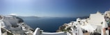 A picturesque coastal scene featuring white-washed buildings with blue domes perched on cliffs overlooking the deep blue Aegean Sea. The sky is clear with a gentle haze on the horizon. A few people can be seen enjoying the view.