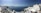 A picturesque coastal scene featuring white-washed buildings with blue domes perched on cliffs overlooking the deep blue Aegean Sea. The sky is clear with a gentle haze on the horizon. A few people can be seen enjoying the view.