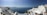 A picturesque coastal scene featuring white-washed buildings with blue domes perched on cliffs overlooking the deep blue Aegean Sea. The sky is clear with a gentle haze on the horizon. A few people can be seen enjoying the view.