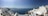 A picturesque coastal scene featuring white-washed buildings with blue domes perched on cliffs overlooking the deep blue Aegean Sea. The sky is clear with a gentle haze on the horizon. A few people can be seen enjoying the view.