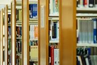 A series of wooden bookshelves filled with a variety of books organized vertically. The image captures a library setting with clear signs of academic or educational content, given the visible titles and categorization of the books.