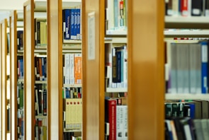 A series of wooden bookshelves filled with a variety of books organized vertically. The image captures a library setting with clear signs of academic or educational content, given the visible titles and categorization of the books.