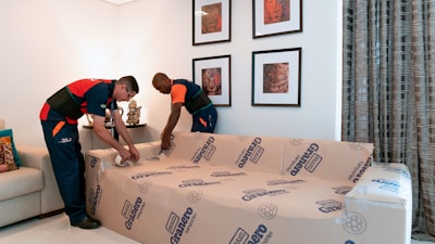 Two individuals in uniform are wrapping a sofa in protective material in a living room. The room features framed artwork on the wall, a neutral-colored sofa to the side, decorative cushions, and patterned curtains.