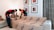 Two individuals in uniform are wrapping a sofa in protective material in a living room. The room features framed artwork on the wall, a neutral-colored sofa to the side, decorative cushions, and patterned curtains.