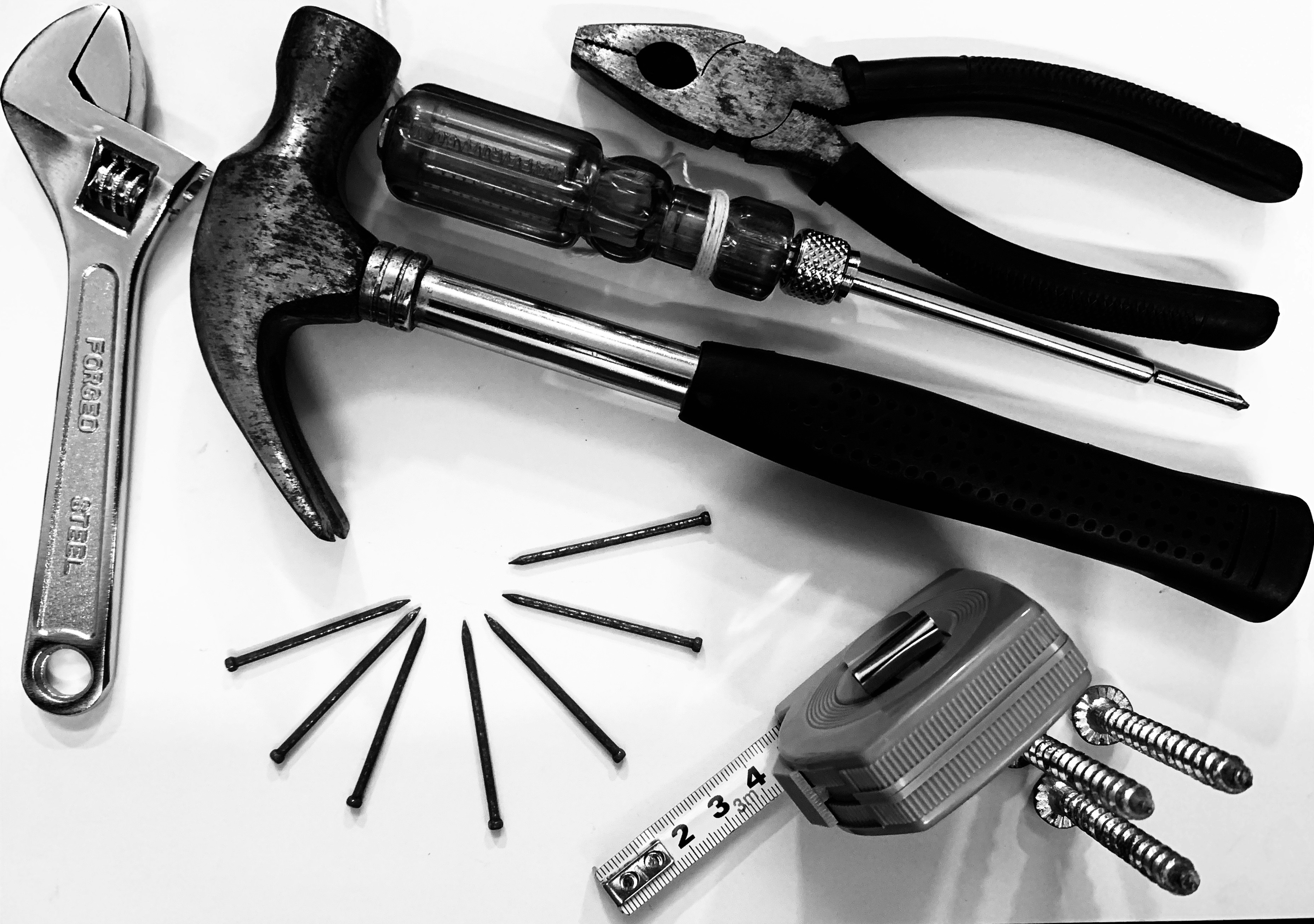 A collection of tools including pliers, a hammer, a screwdriver, an adjustable wrench, several nails, and a tape measure. The tools are arranged on a white surface, showcasing their metallic and rubber textures.