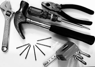 A collection of tools including pliers, a hammer, a screwdriver, an adjustable wrench, several nails, and a tape measure. The tools are arranged on a white surface, showcasing their metallic and rubber textures.