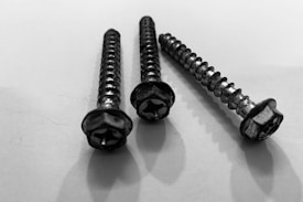 Three metal screws are arranged on a flat surface, casting shadows. The screws have helical ridges along their bodies and hexagonal heads, appearing worn with slight rust.