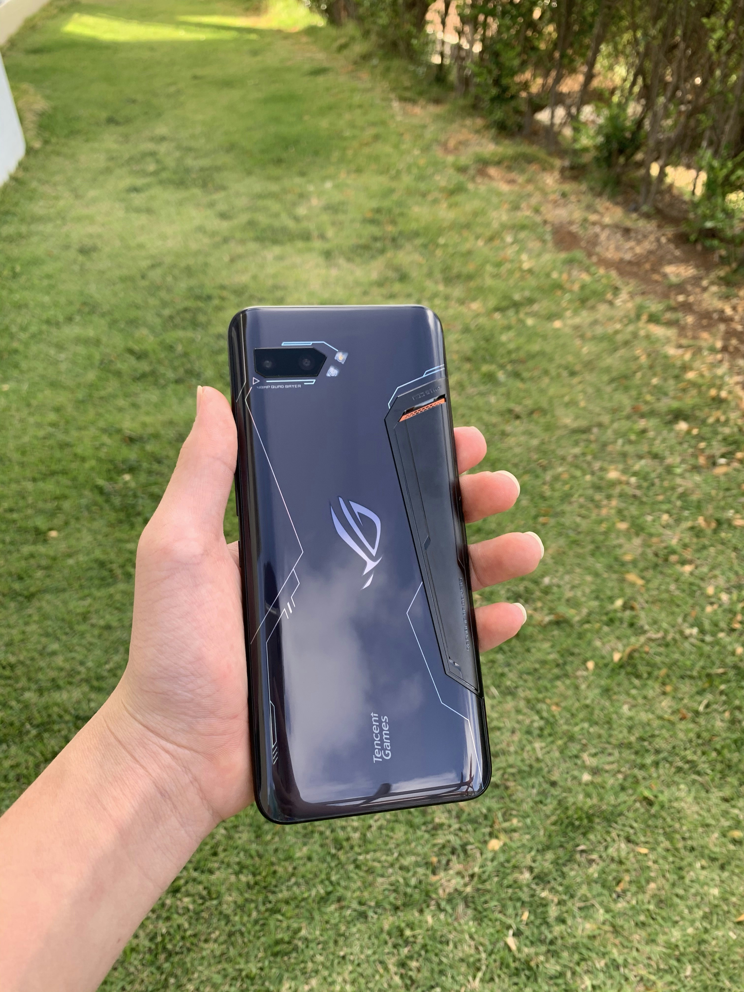 A hand holding a sleek, dark-colored smartphone with geometric patterns and the text 'Tencent Games' on the back cover. The background features a grassy area with some trees or bushes on the side.