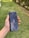 A hand holding a sleek, dark-colored smartphone with geometric patterns and the text 'Tencent Games' on the back cover. The background features a grassy area with some trees or bushes on the side.
