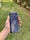 A hand holding a sleek, dark-colored smartphone with geometric patterns and the text 'Tencent Games' on the back cover. The background features a grassy area with some trees or bushes on the side.