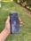 A hand holding a sleek, dark-colored smartphone with geometric patterns and the text 'Tencent Games' on the back cover. The background features a grassy area with some trees or bushes on the side.