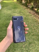 A hand holding a sleek, dark-colored smartphone with geometric patterns and the text 'Tencent Games' on the back cover. The background features a grassy area with some trees or bushes on the side.