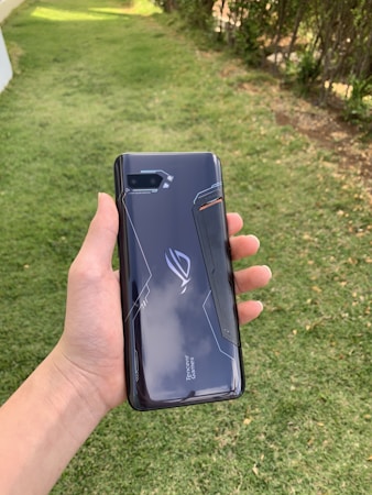 A hand holding a sleek, dark-colored smartphone with geometric patterns and the text 'Tencent Games' on the back cover. The background features a grassy area with some trees or bushes on the side.