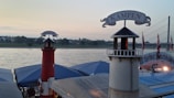 Two decorative lighthouse structures with signs reading 'HORNUM' and 'KAMPEN' are situated by a riverside. The lighthouses are red and white in color, standing on what appears to be a dock or pier area. In the background, a calm river stretches across, with a grassy bank and residential buildings visible. The sky is overcast with a soft evening or morning light, and a bridge can be seen on the right side.