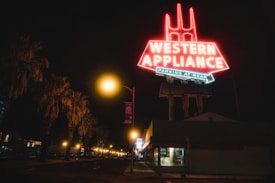 A brightly lit neon sign reading 'Western Appliance' dominates the night scene. The sign is elevated on tall supports and has additional text below indicating parking at the rear. The street is lined with palm trees, and the area is illuminated by streetlights along a seemingly quiet road. A building beneath the sign appears closed, with dimly lit windows.